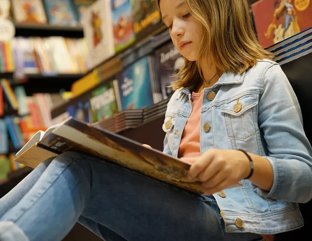 Une petite fille lit une BD assise par-terre contre un étagère